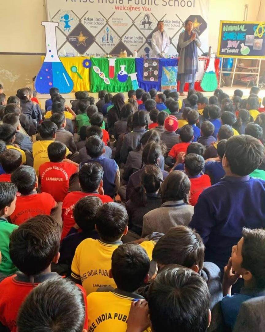 Students seated in an auditorium for a National Science Day presentation with science-themed stage decorations